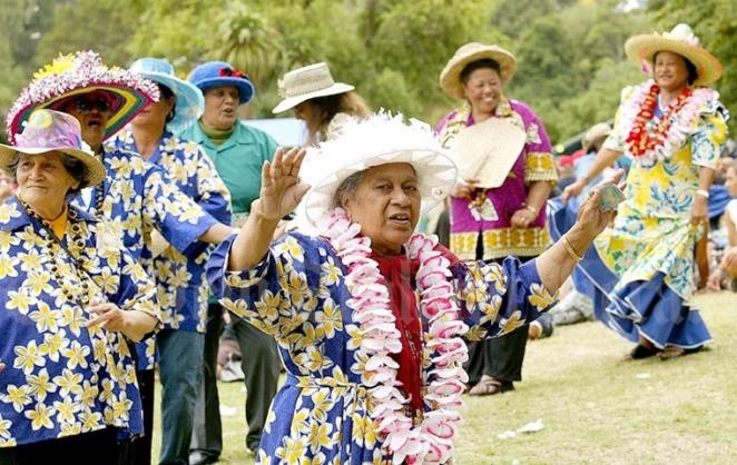 A group of Pacific Island women wearing hats, leis, and colourful shirts dance in a field