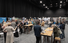 People interacting at a trade booth area, viewing displays and engaging with representatives. The space is lively, with various booths showcasing products and services.