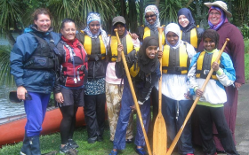 A group of women stand in front of a river, smiling. Some are holding oars
